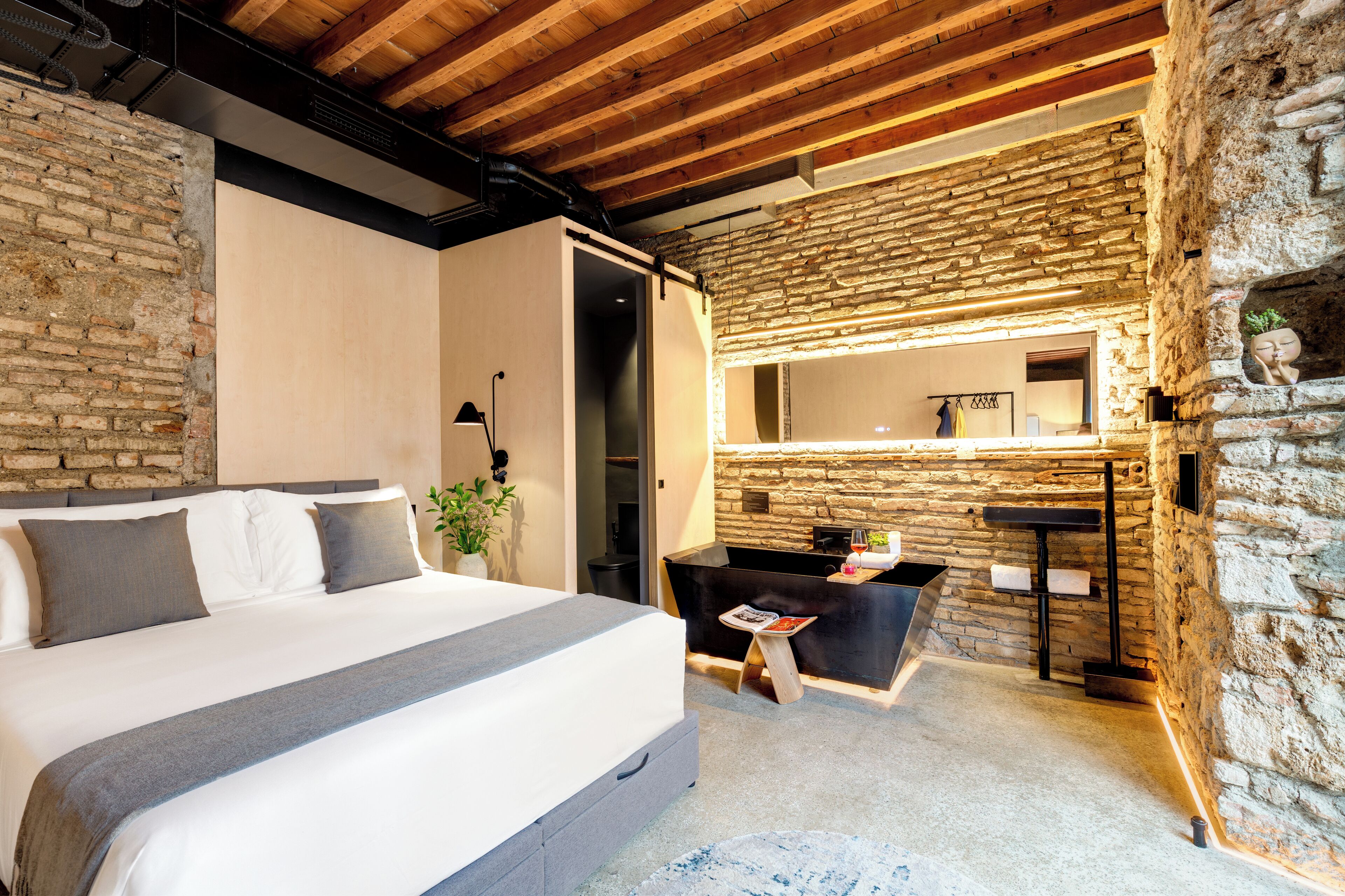 A bedroom with a large bed, a stone wall, a wooden ceiling, and a small table with a magazine.