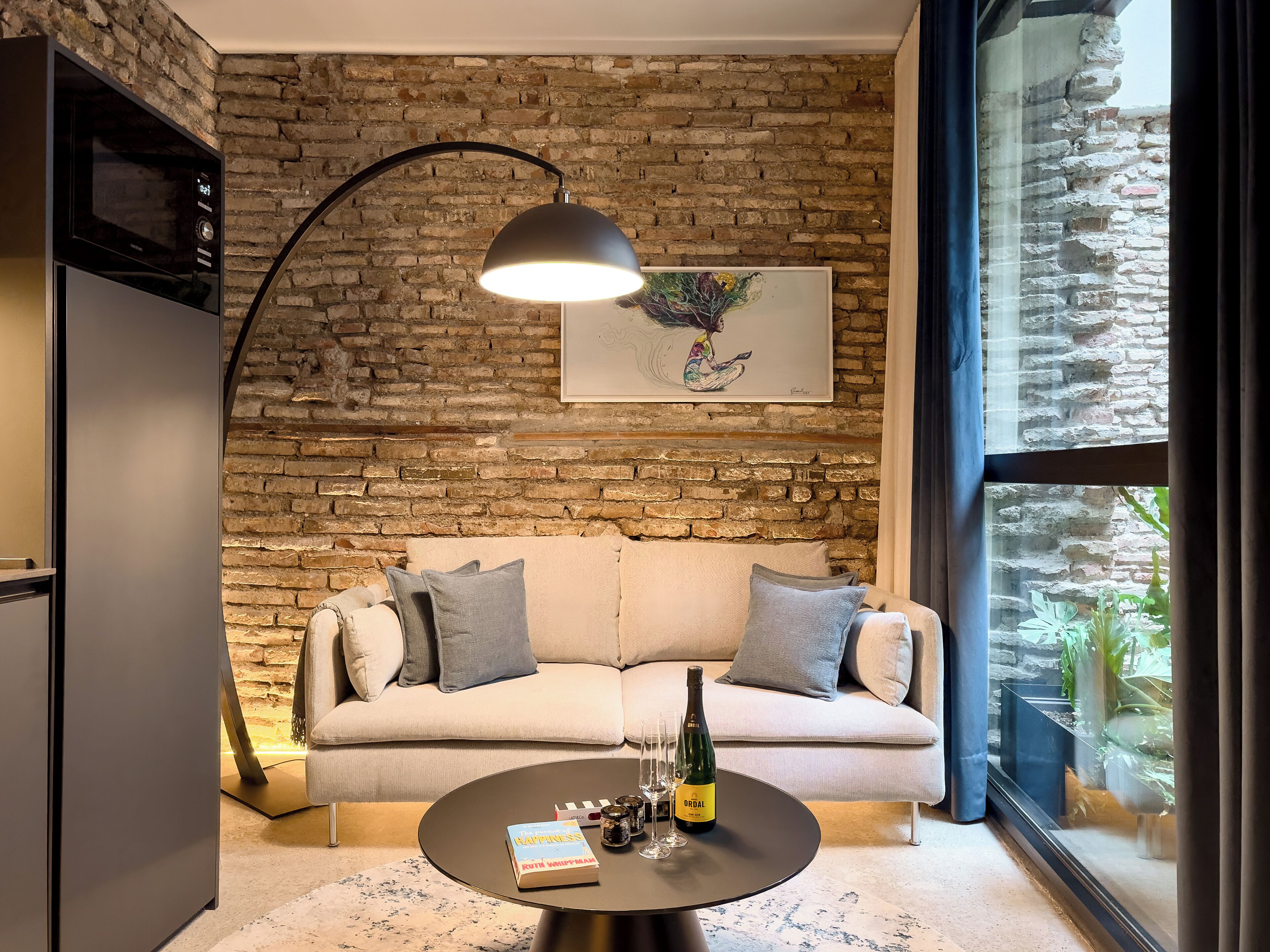 Een gezellige woonkamer met een bakstenen muur, een bank, een bijzettafeltje met een boek en een fles, en een hanglamp.