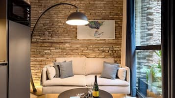 Una sala de estar acogedora con una pared de ladrillo, un sofá, una mesa baja con un libro y una botella, y una lámpara colgante.