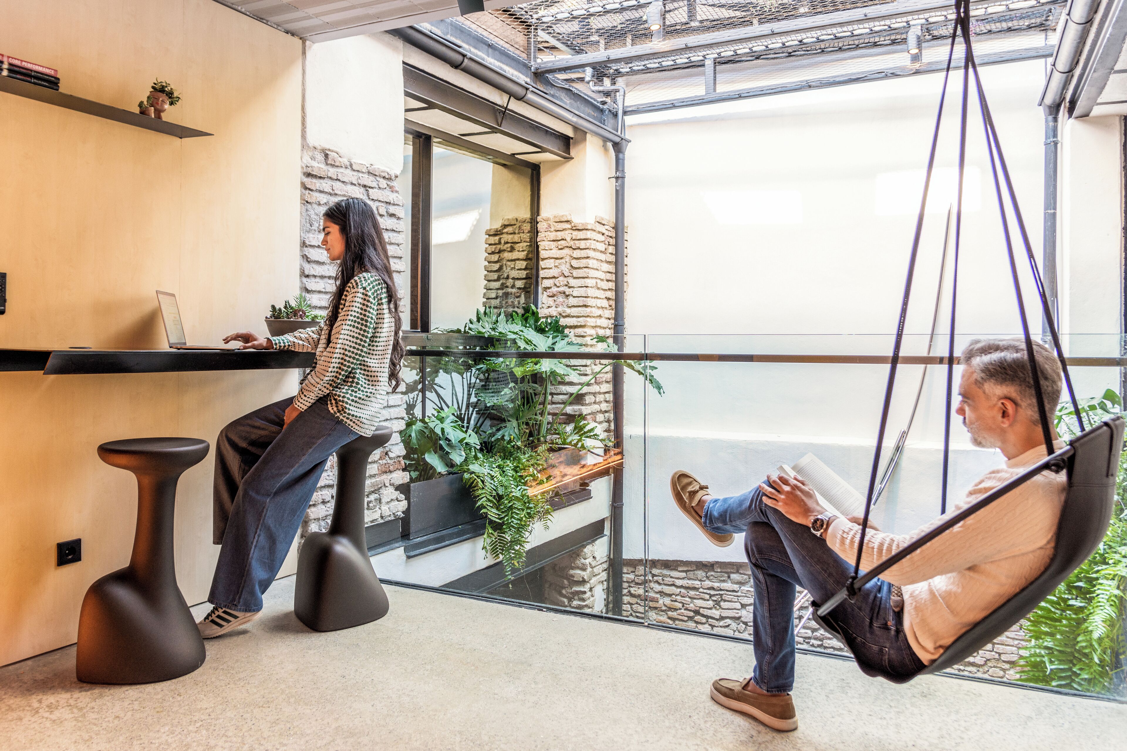 A modern office space with a glass wall, a suspended chair, and a person working at a desk.