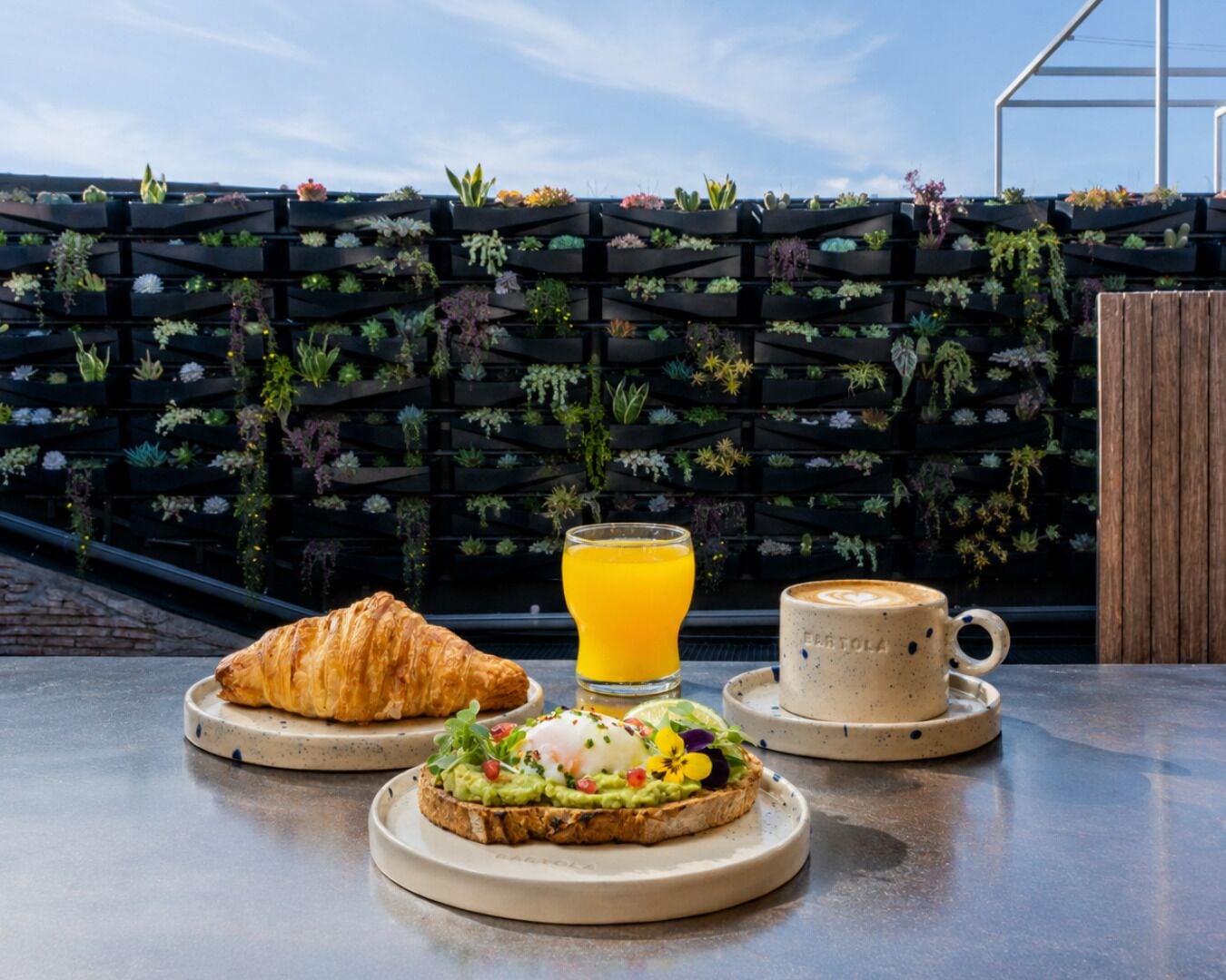 En frukostuppdukning med croissanter, avokadomackor och kaffe på ett bord med utsikt över en vertikal trädgård.