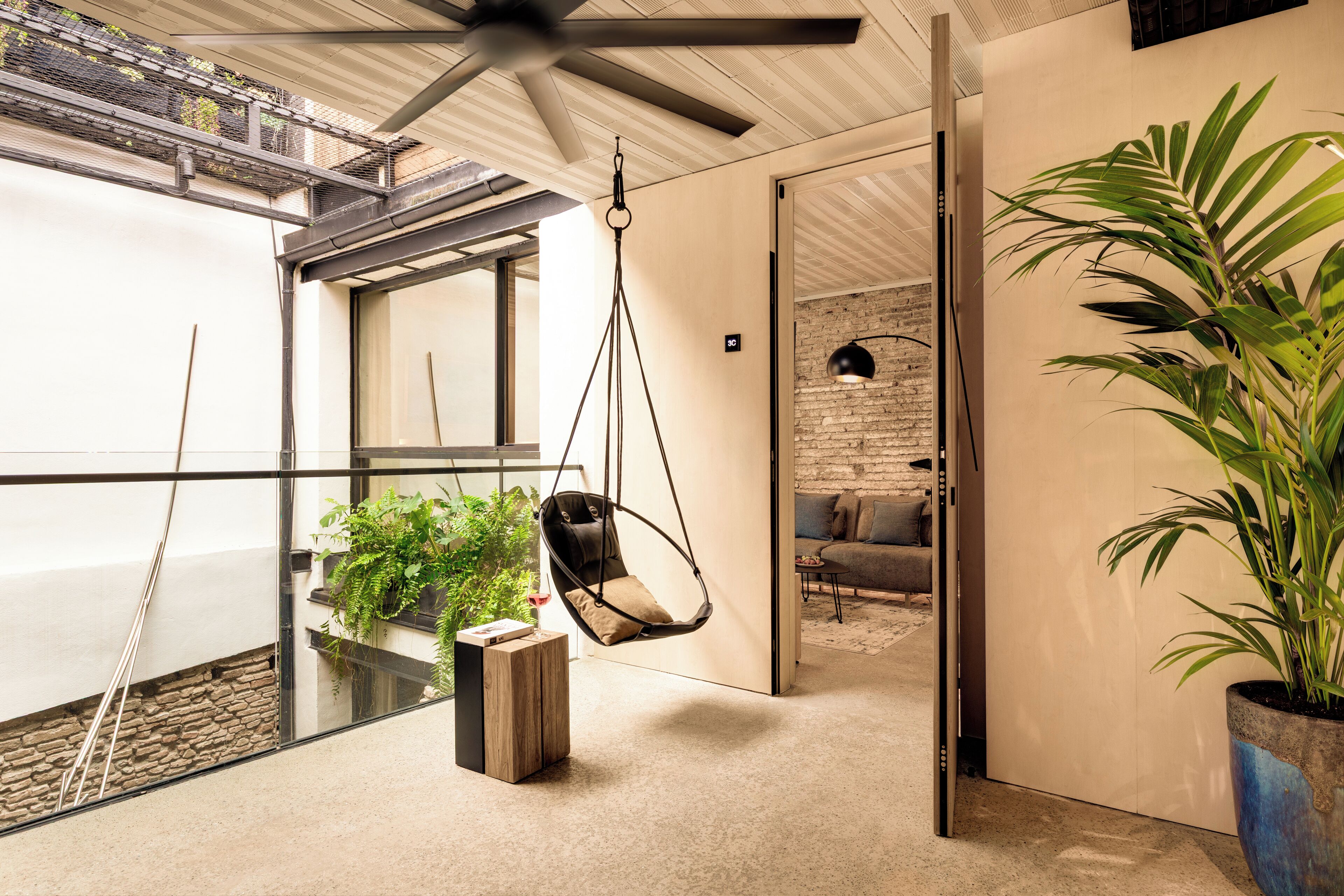Un intérieur moderne avec un fauteuil suspendu, une grande plante en pot et une vue sur un mur de briques.