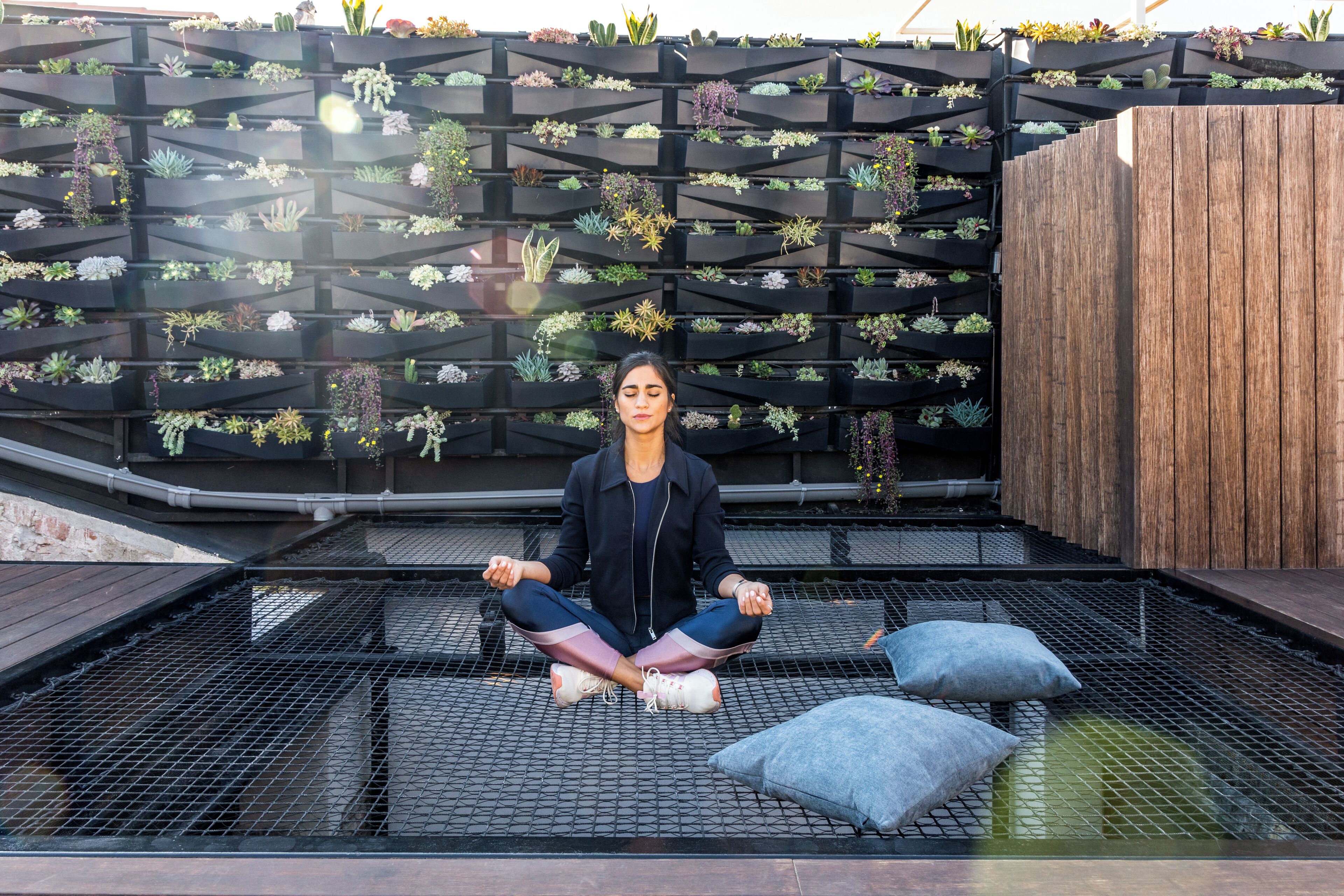 A person meditating on a rooftop with a garden wall behind.