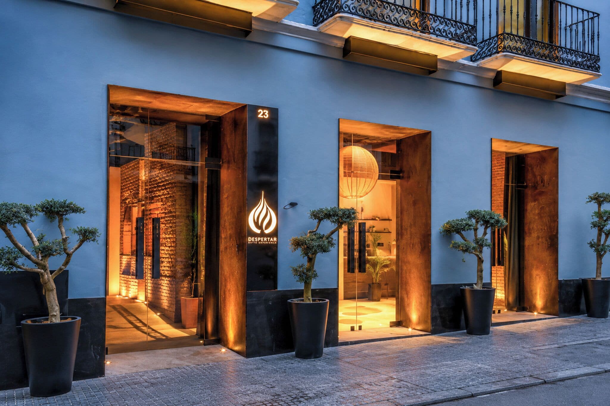Entrada de un hotel con un letrero con el número 23, un logo de protección contra incendios y árboles en macetas.