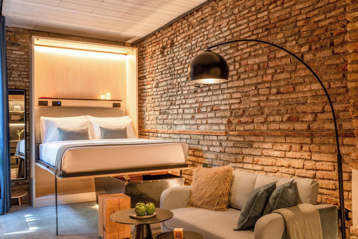 A modern hotel room with a brick wall, a bed, a sofa, and a round table with apples.