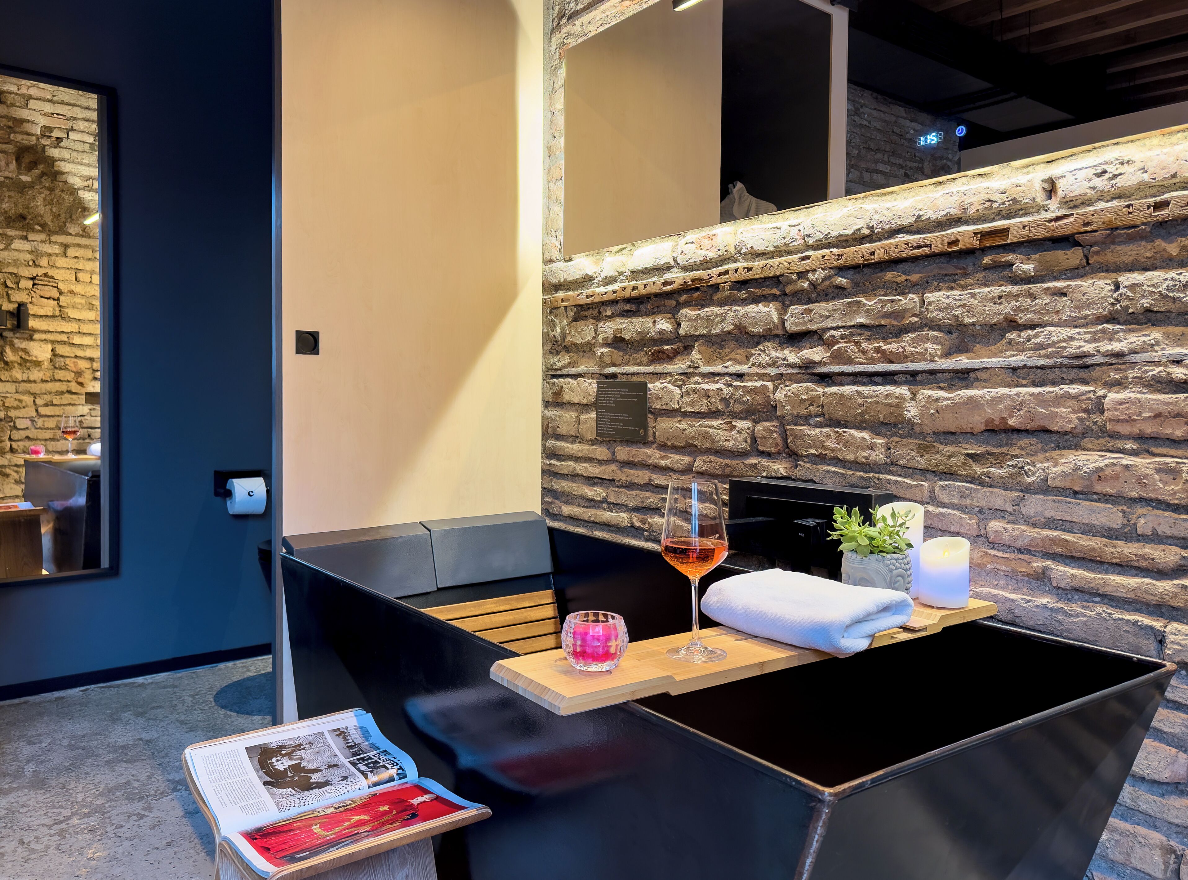 Un área de estar exterior moderna con un muro de piedra como fondo, un banco de madera, una mesa con una copa de vino, una vela y una pequeña planta.