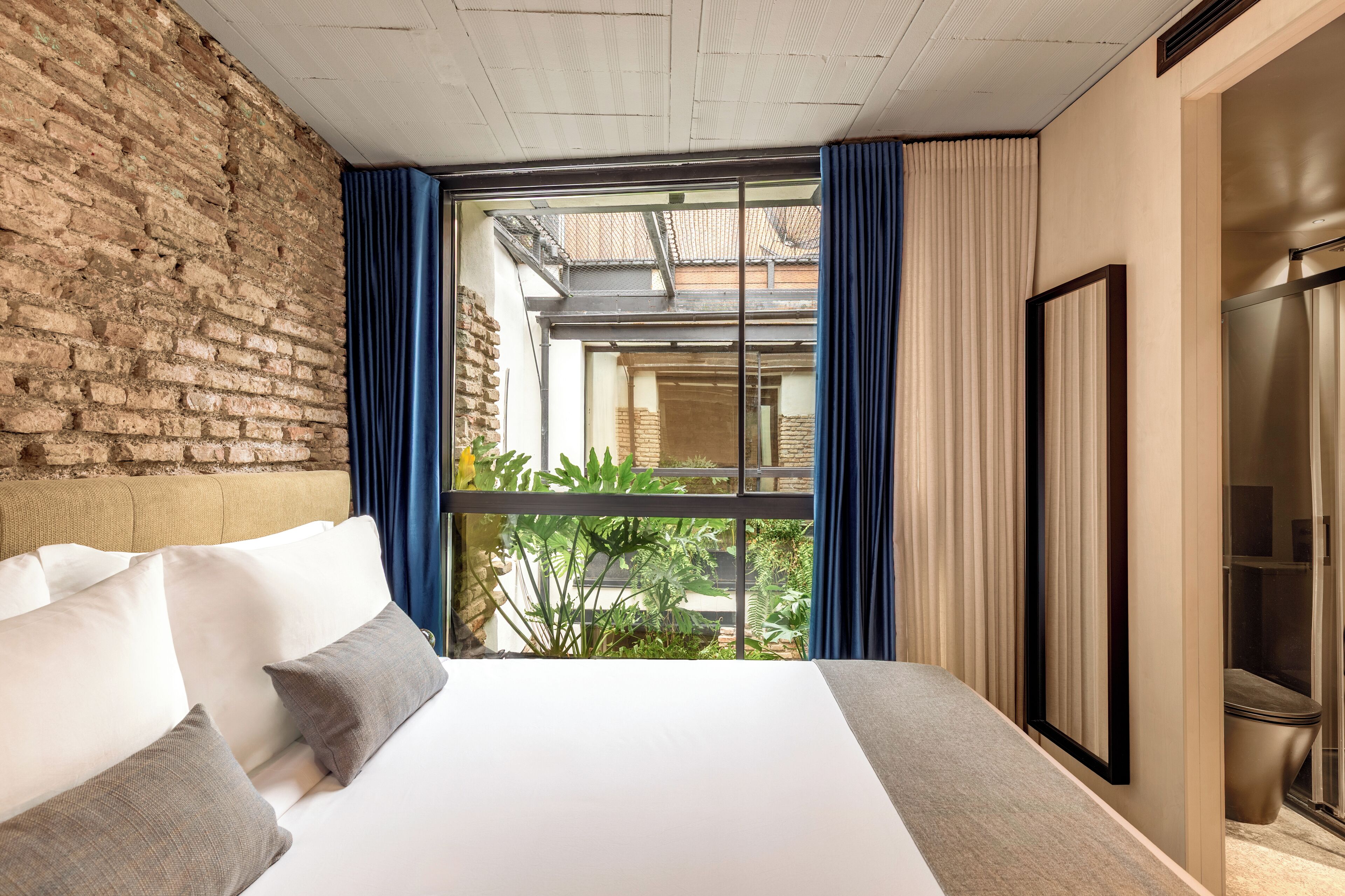 Habitación de hotel con una cama grande, pared de ladrillo, vista al patio interior y una puerta corredera al baño.