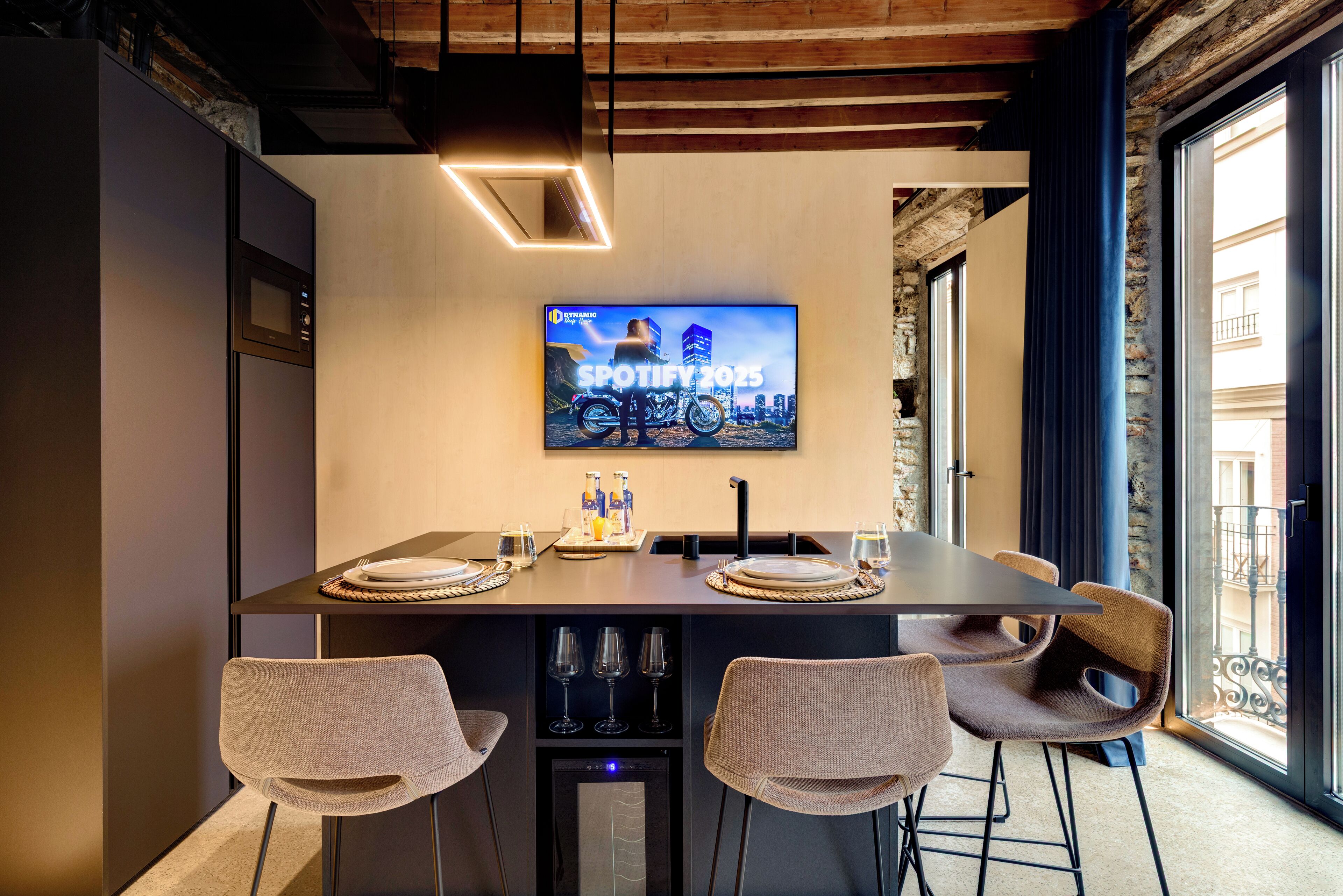 A modern dining area with a long table, four chairs, a television displaying 'SPOTIFY 2025', and a city view through large windows.