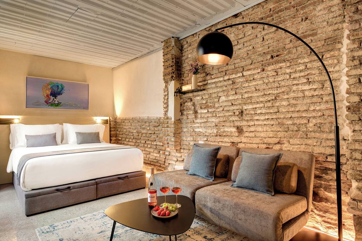A modern hotel room with a brick wall, a bed, a sofa, a coffee table with a bottle and glasses, and a painting on the wall.