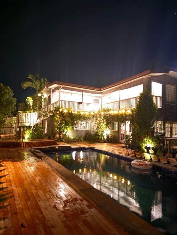 Une maison à deux étages avec une piscine, entourée de verdure et illuminée par des guirlandes lumineuses.