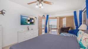 Ett sovrum med en säng, en tv som är monterad på väggen, en takfläkt och utsikt över havet genom en öppen dörr.