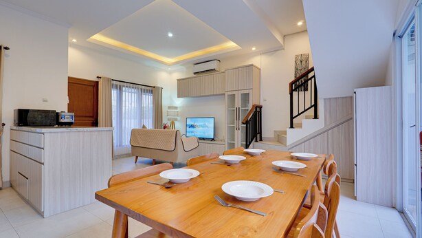 A modern dining area with a wooden table set for six, a kitchen with a built-in oven, and a living area with a TV and seating. - Villa Taman Seminyak 52 - Seminyak (Seminyak)