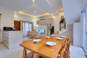A modern dining area with a wooden table set for six, a kitchen with a built-in oven, and a living area with a TV and seating. - Villa Taman Seminyak 52 - Seminyak (Seminyak)