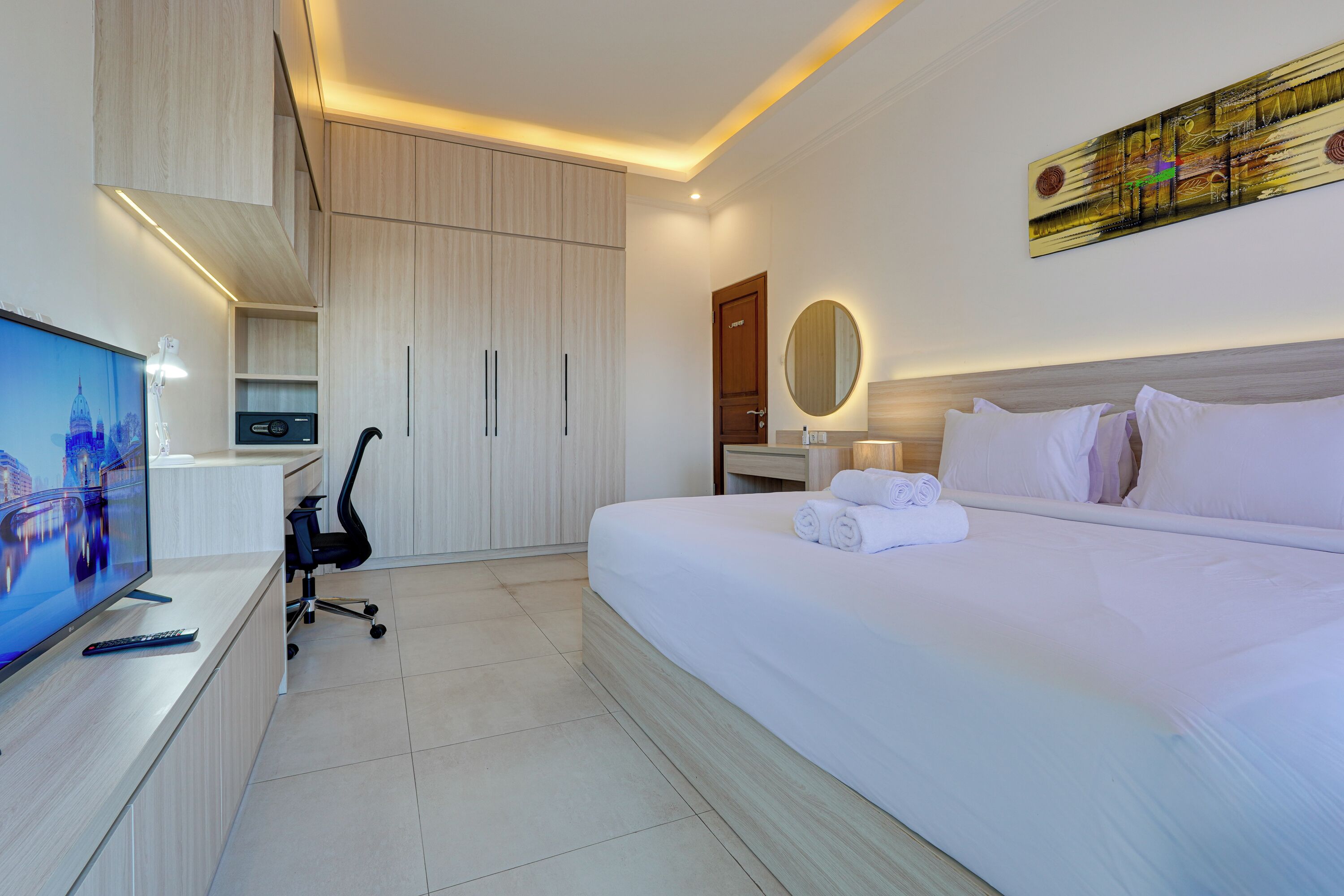 A hotel room with a bed, a desk with a computer and remote control, a chair, a TV, a mirror, and a wall art piece.