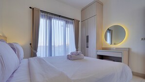 A neatly made bed with white linens and rolled towels, a bedside table with a lamp, a round mirror with a light, and a window with sheer curtains. - Villa Taman Seminyak 52 - Seminyak (Seminyak)