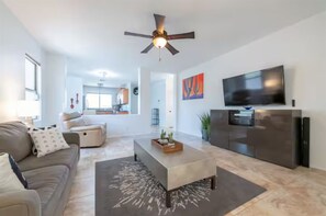 A modern living room with a sofa, coffee table, TV, and kitchen area in the background. -  Optional heated pool- 3-bedroom house in peaceful Surprise with AC (Surprise)