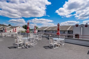 Eine Dachterrasse mit weißen Stühlen und roten Streifen-Schirmen, mit Blick auf eine Küstenregion mit Gebäuden und blauem Himmel.