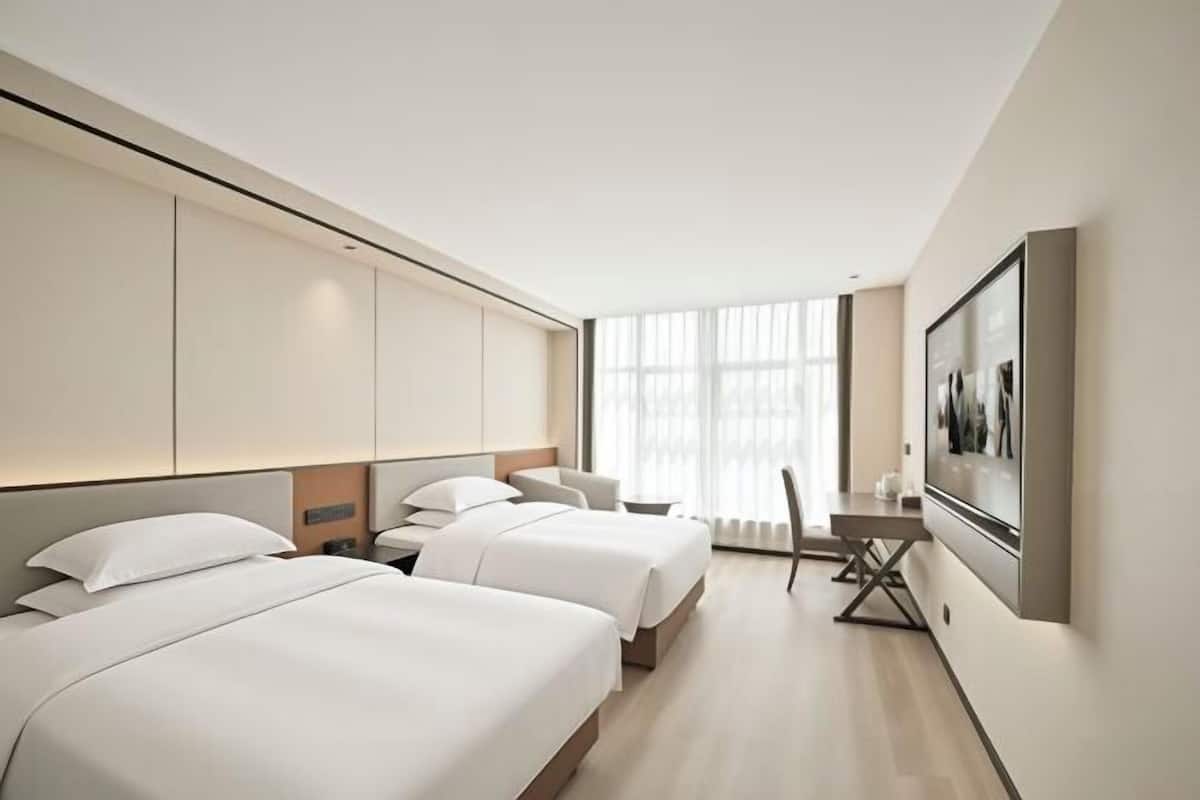 A hotel room with two beds, a desk, and a large window with sheer curtains.