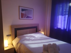 A hotel room with a bed, a nightstand with a lamp, a wall-mounted painting, and a window with curtains.