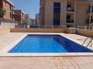 Una piscina rectangular con plataforma empaveda, rodeada por una cerca y edificios adyacentes.
