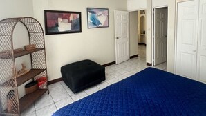 Une chambre avec un grand lit, une étagère en bois avec des boîtes de rangement, deux tableaux encadrés au mur et une porte blanche donnant sur une autre pièce.