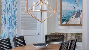 Uma sala de jantar com mesa de madeira, cadeiras pretas, um lustre trançado, um quadro de um navio no mar e uma parede azul abstrata.