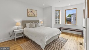 Une chambre à coucher avec un grand lit, des lampes de chevet, un banc, un tableau et une fenêtre donnant sur des immeubles.