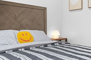 A neatly made bed with a striped bedspread, a pillow with a smiley face, a bedside lamp, and a wooden headboard. - Slice of the good Life: Concord A8 (Peoria)