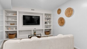 Een woonkamer met een flatscreen televisie, witte schappen, rieten manden en een witte bank.