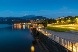 A waterfront promenade with a stone wall, a bridge, and a view of a town and mountains in the background. - Bee Apartment: Luino Lakeside With Outdoor Dining (Luino)