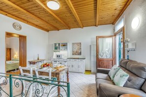 A cozy living area with a wooden ceiling, a dining table with chairs, a sofa with cushions, a kitchen area with cabinets, and a small dining table with food and drinks. - Butterfly Apartment: Lake Maggiore Luino Apartment With Balcony (Luino)
