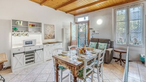 A dining area with a wooden table and chairs, a white cabinet with shelves, a microwave, and a window with decorative curtains. - Butterfly Apartment: Lake Maggiore Luino Apartment With Balcony (Luino)