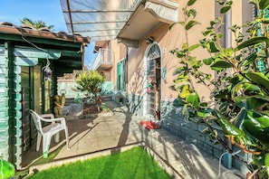 Un patio con césped verde, una silla de plástico blanca, un pequeño árbol y una pared de piedra.