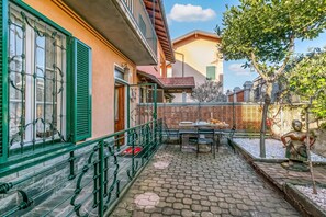 A courtyard with a table and chairs, a statue, and a building with green shutters and a balcony. - Butterfly Apartment: Lake Maggiore Luino Apartment With Balcony (Luino)