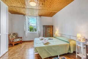 A bedroom with a wooden ceiling, a bed with a green quilt, a wooden chair, a window with curtains, and a nightstand with a lamp. - Butterfly Apartment: Lake Maggiore Luino Apartment With Balcony (Luino)