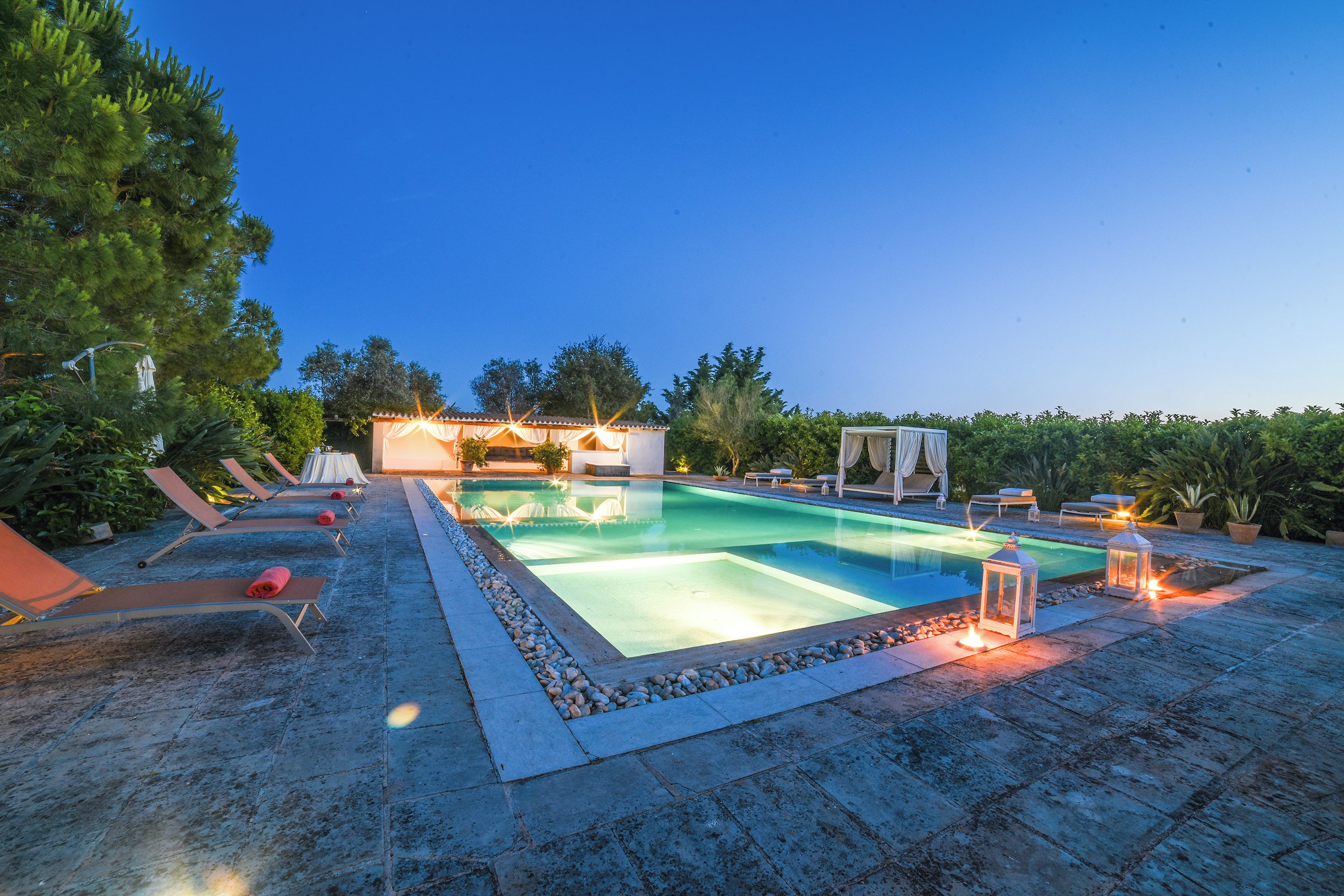 A rectangular pool with lounge chairs and umbrellas, surrounded by a stone deck and greenery.