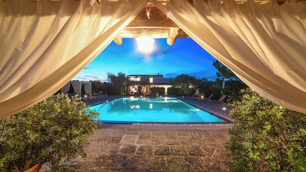 Piscina com vista para uma casa ao fundo, cercada por vasos de plantas e uma pérgula com cortinas.