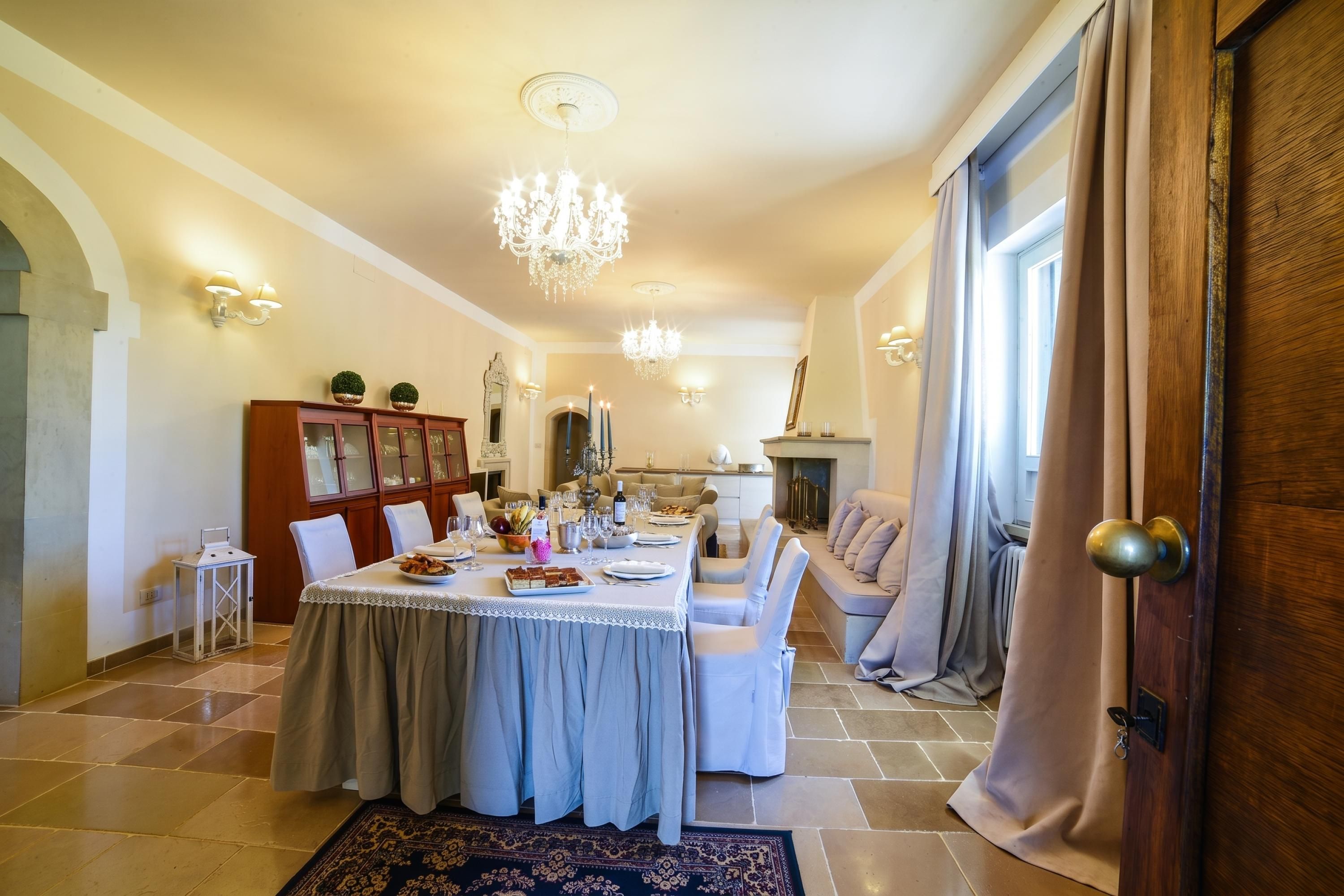 A dining area with a long table, chairs, and a chandelier. There is a sofa and a fireplace in the background.