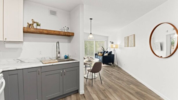 Eine moderne Küche mit grauen Schränken, einer Marmorarbeitsplatte und einem Holzregal. Es gibt einen Essbereich mit Tisch und Stühlen sowie ein Wohnzimmer mit Sofa und Lampe.