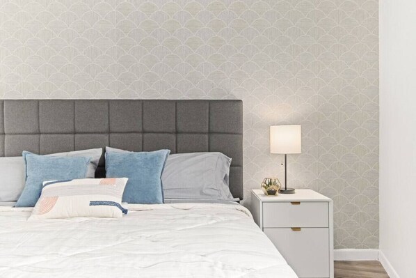 A neatly made bed with a grey headboard, white bedding, and blue pillows. A bedside table with a lamp and a decorative item is visible. - Slice of the Good Life: Concord C2 (Peoria)