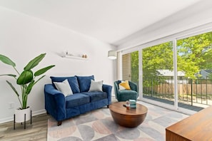 A modern living room with a blue sofa, a round coffee table, and a green armchair. There is a large window providing a view of the outdoors. - Slice of the Good Life: Concord B6 (Peoria)
