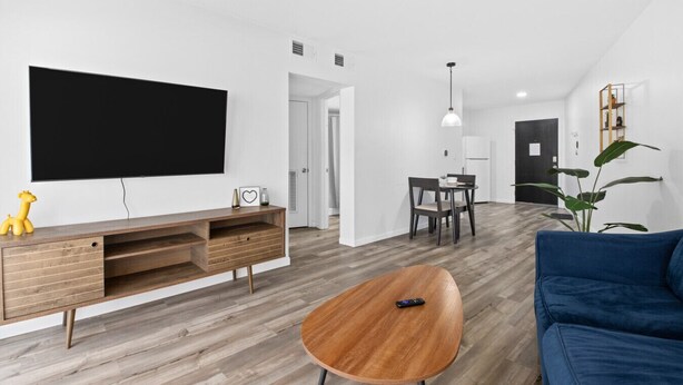 Una sala moderna con televisor de pantalla plana, mueble de entretenimiento de madera, mesa de centro redonda, sofá azul, mesa de comedor con sillas y una planta en maceta.