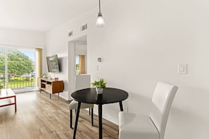 Ein modernes Wohnzimmer mit einem runden schwarzen Tisch, weißen Stühlen, einem weißen Sofa, einem Fernseher an der Wand und einem Balkon mit Blick auf Grünflächen und einen Zaun.