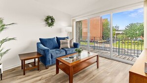 A modern living room with a blue sofa, a wooden coffee table, and a large sliding glass door leading to a balcony with a view of the street and greenery. - Concord C1 · Slice of the Good Life: Concord C1 (Peoria)
