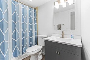 Un baño con inodoro blanco, mueble gris, lavabo blanco y cortina de ducha azul estampada.