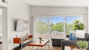 Een moderne woonkamer met een bank, salontafel en televisie. Er is een balkon met uitzicht op groen en een huis.