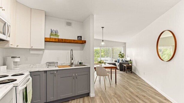 Een moderne keuken met grijze kastjes, een wit aanrechtblad en een houten plank boven de spoelbak. Er is een zithoek met een tafel en stoelen, en een grote spiegel aan de muur.