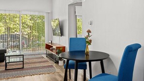 A modern living room with a round table, blue chairs, a glass coffee table, a TV on the wall, and a view of greenery outside. - Concord C6 · Slice of the Good Life: Concord C6 (Peoria)