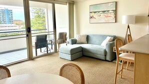 Ein modernes Wohnzimmer mit grauem Sofa, einem Esstisch, Korbstühlen und einem großen Fenster mit Blick auf Gebäude und Bäume.