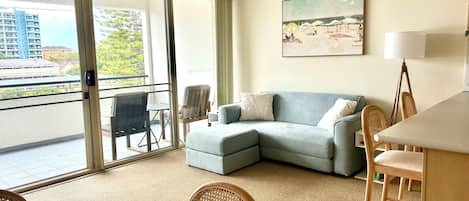 Una sala de estar moderna con un sofá gris, una mesa de comedor, sillas de mimbre y un ventanal con vistas a edificios y árboles.